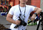 A young male journalist in a white tshirt with a camera around his neck leans against a tripod in a village street.